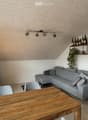 Modern living room with grey sectional sofa, wooden dining table, chairs, wine rack, shelves with bottles and plants, and a floor lamp.