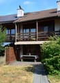 Two-story house with a tiled roof, solar panels, and a wooden balcony. The front yard includes a wooden fence, a small patio with a table and chairs, and lush greenery.