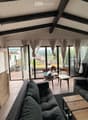 Living room with large windows and sliding doors leading to a patio, featuring a dark green sofa, a coffee table, and several chairs.