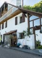 Two-story house with white walls, wooden accents, large windows, and a balcony on the upper level. The house features a stone chimney, a small garden area with potted plants, and a paved driveway.