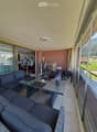 Balcony with a grey sofa, wicker table, chairs, flower pots, and a barbecue. Large windows offer a scenic view of the surrounding landscape.