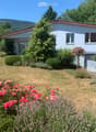 A single-story house with a red roof and light blue walls, surrounded by a well-maintained garden with various flowering plants, shrubs, and trees. The front yard features a gravel pathway leading to the house entrance.