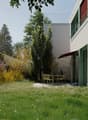 A modern garden area with a lawn, small trees, and a patio with outdoor furniture near a contemporary building.