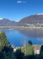Panoramic view of a lake surrounded by snow-capped mountains, with trees and a small settlement visible on the shore.