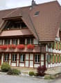Two-story traditional house with red-tiled roof, white and brown facade, balcony with red flowers, green shutters, and small garden area.