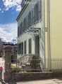 Building. A two-story building with light green walls, multiple windows with shutters, a decorative wrought iron balcony, and a small garden area behind a metal fence.
