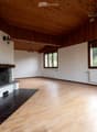 Spacious, unfurnished room with wooden floors, white walls, large windows, and a stone fireplace. The room features exposed wooden beams on the ceiling and radiators along the walls.
