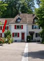 Two-story house with red shutters, white walls, and a tiled roof. The house features a driveway, garden, and is surrounded by trees and greenery.