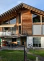 Two-story wooden house with large windows, balcony, outdoor seating area, and garden.