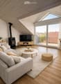 Modern living room with wooden floors, slanted wooden ceiling, large windows, balcony access, sofa, coffee table, television, fireplace, and decorative items.