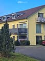 Two-story yellow house with red-tiled roof, multiple windows, balconies, and a driveway with a parked car.