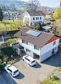 Aerial view of a two-story house with a garage, driveway, and surrounding greenery. Two cars are parked in the driveway, and there are additional houses and greenery in the background.