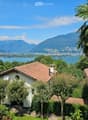 A serene garden view with a small white house, surrounded by lush greenery and palm trees, overlooking a lake and distant mountains.