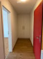 A narrow hallway with wooden flooring, light-colored walls, and a built-in closet with double doors. The hallway features a red accent wall on one side and a partially open door leading to another room.