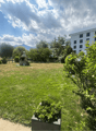 A spacious, well-maintained garden area with green grass, trees, shrubs, and a small stone fireplace in the background. The area is bordered by a modern building and features a clear, partly cloudy sky.