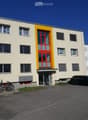 Two-story building with light beige walls, red and yellow entrance door, multiple windows with blinds, and a small parking area with bicycles and a car.