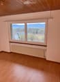 Empty room with wooden flooring, large windows offering a scenic mountain and lake view, radiator, and wooden ceiling beams.