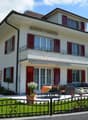 Two-story house with white exterior, red shutters, and a balcony on the upper floor. The front yard features a well-maintained lawn, a black wrought-iron fence, and outdoor seating including a sofa, coffee table, and dining table with chairs.