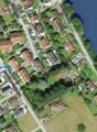 Aerial view of a residential area with multiple houses, gardens, and surrounding fields. The houses are clustered along streets, with some near a body of water.