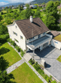Aerial view of a two-story house with a brown tiled roof, white walls, and a spacious balcony. The property is surrounded by a well-maintained lawn and trees, with neighboring houses visible in the background.