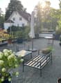 A well-maintained garden area with gravel ground, potted plants, a metal bench, a small table, and chairs, with an umbrella and surrounding greenery.