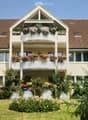 Two-story house with a brown tiled roof, white exterior, multiple windows, and balconies adorned with flower boxes and greenery.
