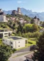 House with solar panels on the roof, raised ground floor, surrounded by trees and plants, with a view of mountains in the background.