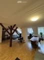 Open-plan living and dining area with wooden floors, exposed wooden beams, large windows, sofa, coffee table, dining table, chairs, and a plant.