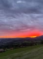 A panoramic view of a rural landscape at sunset, featuring rolling green hills, scattered houses, a body of water, and distant mountains under a dramatic sky.