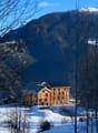 A modern wooden house with a dark roof, situated in a snowy mountainous landscape with bare trees in the foreground.