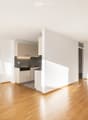 Open-plan unfurnished room with light wooden flooring, white walls, and a small kitchen area featuring cabinets, sink, and dishwasher.