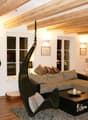 Living room with exposed wooden beams, large windows, a grey sectional sofa, a black wicker hanging chair, a coffee table, and a dining area with chairs and a table.