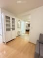 A modern, minimalist living room with light wooden flooring, white walls, a grey sofa, a white cabinet with glass doors, a small white side table, and a mirror on the wall. The room is connected to another space with a bed visible in the background.
