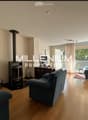 Spacious living room with wooden flooring, fireplace, large windows, and balcony access. Features a blue sofa, armchair, and small table.
