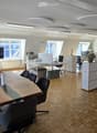 Bright, modern office space with wooden flooring, large windows, a long wooden conference table with chairs, a TV screen, desks, cabinets, and decorative plants.