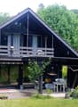 Two-story house with a dark wooden exterior, large windows, and a balcony on the upper level. The surrounding area includes a well-maintained garden with flowering trees and shrubs.
