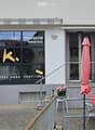 Facade of a commercial building with large windows, signage for "Konfi Manufaktur Markus Kunz" and "LA VIE EST DURE SANS CONFITURE", outdoor seating area with tables and chairs, and a yellow post box.