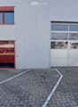 Modern building with a large garage door and a glass entrance. The facade is grey with red trim around the windows and doors. The ground is paved with bricks and marked parking spaces.
