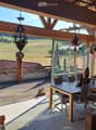 Bright, wooden-framed balcony with large windows offering a scenic countryside view. Features a dining table with chairs, hanging decorations, and potted plants.