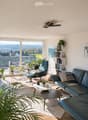Bright living room with large windows and a view, featuring a dark blue sofa, coffee table, armchair, and decorative plants.
