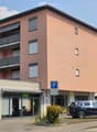 Modern multi-story residential building with pink facade, balconies, and private parking area.