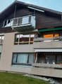 Two-story house with wooden facade, multiple balconies, large windows, and a well-maintained lawn.