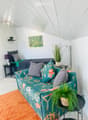 A modern, well-lit room with a green sofa, a bed with dark bedding, a nightstand, a small table with a potted plant, and a picture frame on the wall.