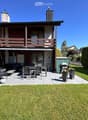 Well-maintained garden with a patio area featuring a sofa, table, chairs, and a grill. The house has a wooden balcony and a well-kept lawn.