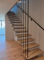 Wooden staircase with black metal balustrade, light wooden steps, and a handrail, set against light-colored walls and wooden flooring.
