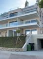 Modern two-story house with large glass balconies, raised ground floor, and garage. The house features a beige facade, multiple large windows, and a landscaped hillside entrance.