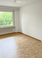 Empty room with wooden flooring, white walls, large window with view of greenery, radiator below window, and a door visible on the left side.