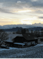 Aerial view of a house with a tiled roof, surrounded by a snow-covered yard and trees, with a scenic mountain backdrop at sunset.