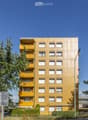 A multi-story residential building with yellow balconies and white-framed windows, surrounded by greenery and a clear blue sky.