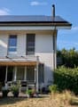 Two-story house with solar panels on the roof, large windows, and a covered patio area with outdoor furniture.